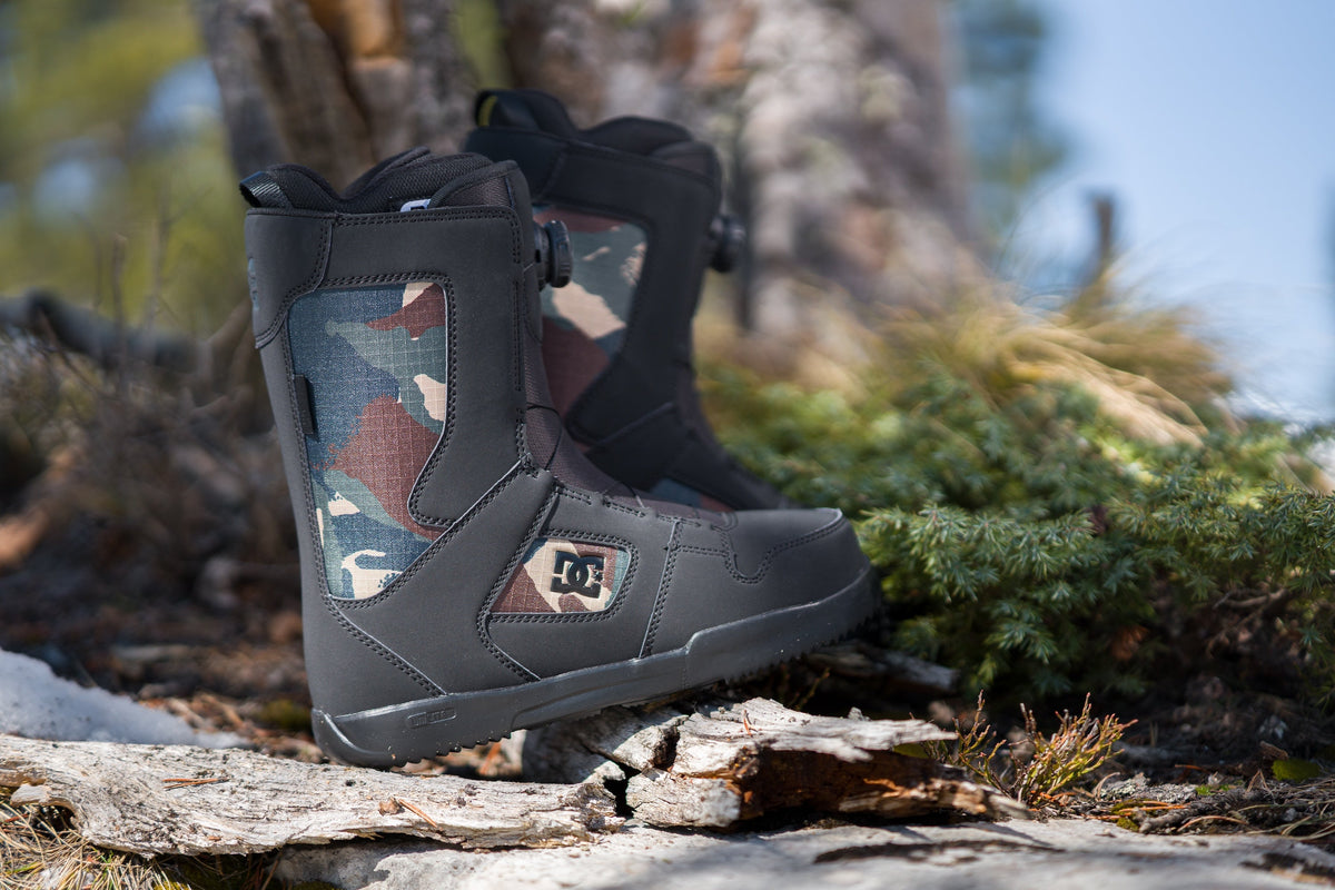 Black DC Shoes Phase BOA snowboard boots with camouflage panels and BOA dial closures, displayed on fallen logs in a wooded outdoor setting.