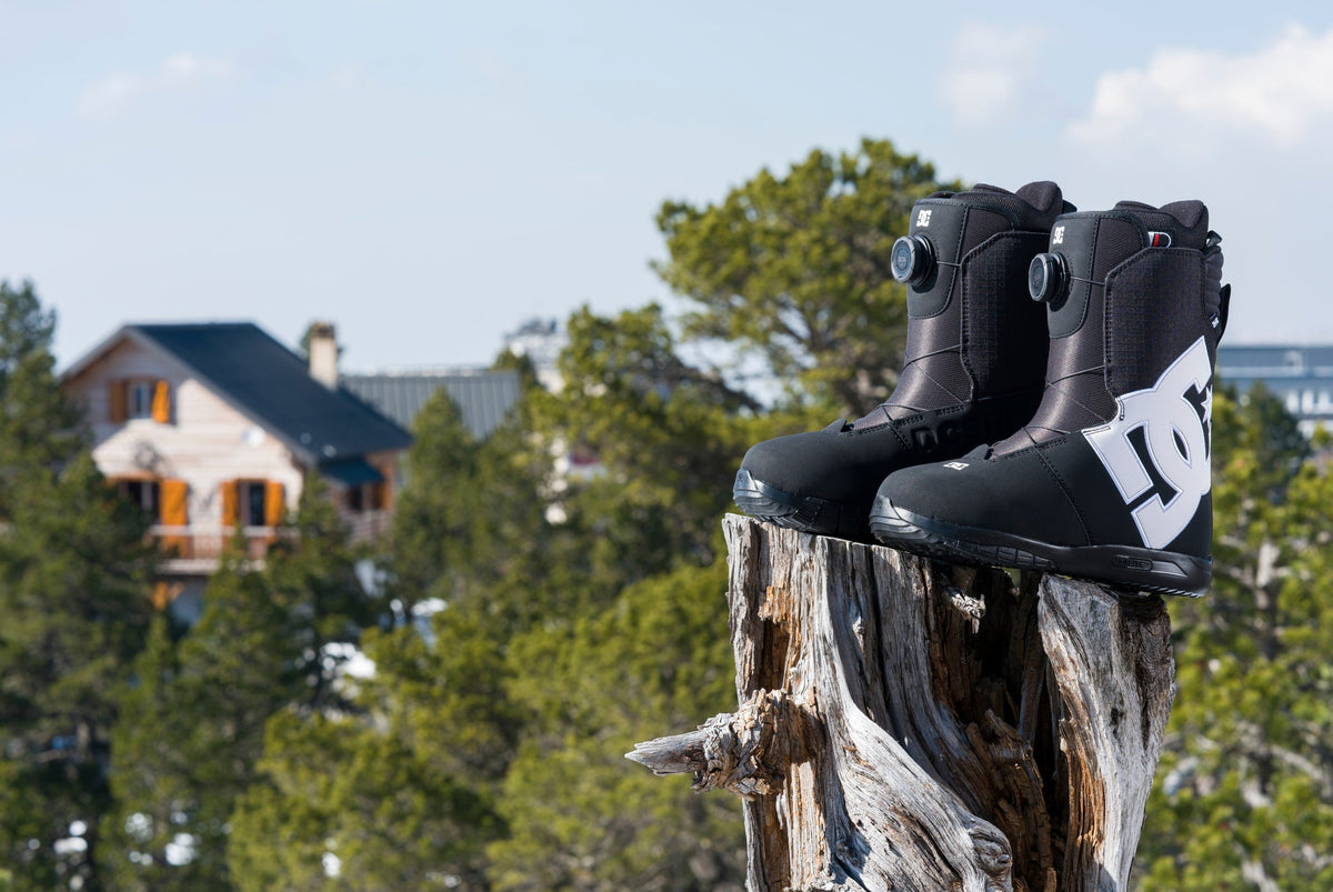 Pair of black DC snowboard boots with BOA closure systems displayed on a tree stump in a mountain setting.