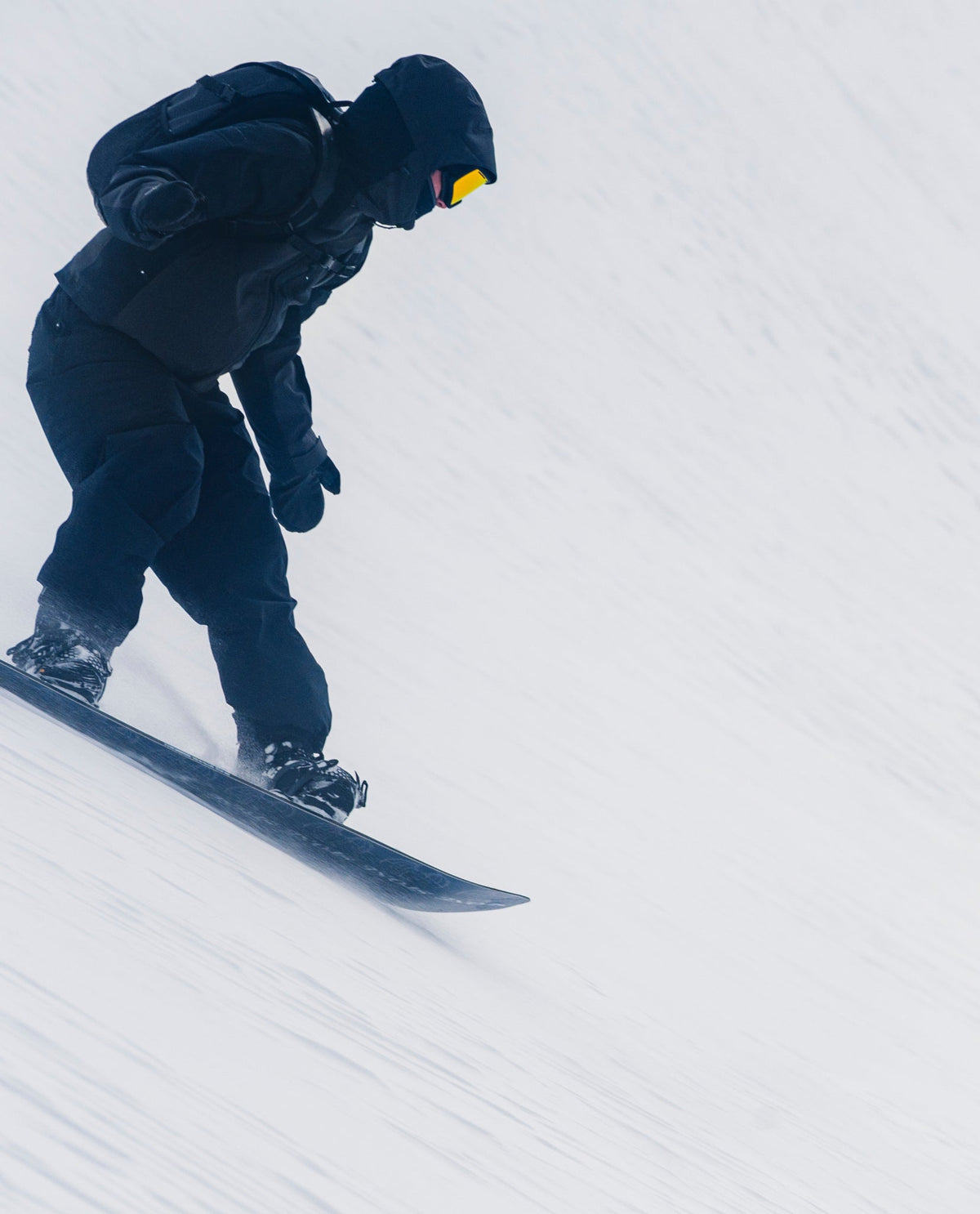 Snowboarder riding down a snowy slope in black outerwear, goggles and a backpack.