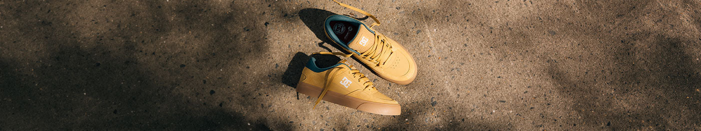 A pair of tan skate shoes with teal interiors and white logos sit on a sunlit, textured concrete surface, casting soft shadows.