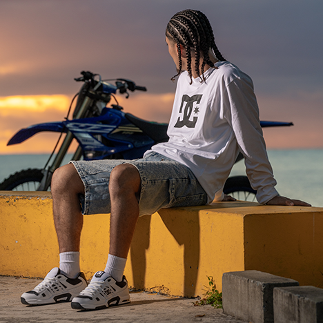 Person wearing a white DC shirt sitting by a motorcycle with a sunset in the background