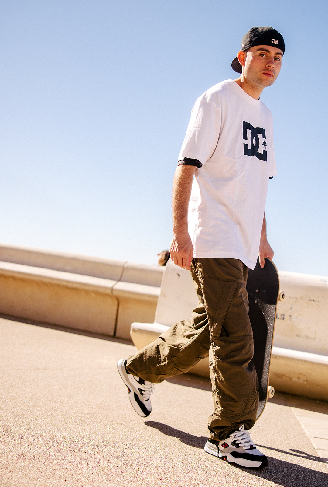Person wearing a white t-shirt with a logo, green pants, and black cap, holding a skateboard on a clear day.