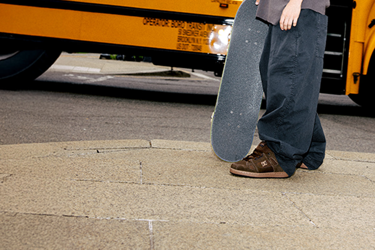 A person standing on a concrete sidewalk in front of a yellow school bus, holding a skateboard upright. The person is wearing loose dark blue pants and brown sneakers.