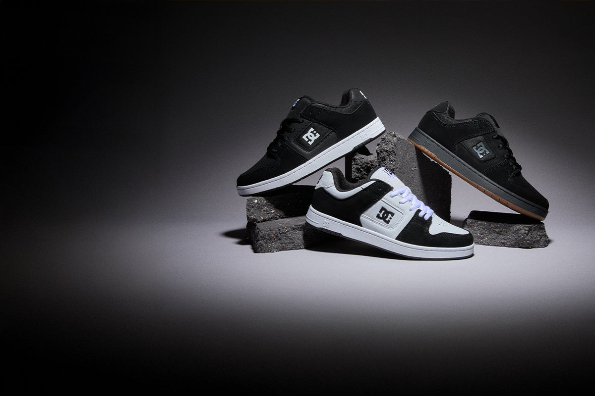 Three DC sneakers displayed on stacked gray blocks against a dark studio background. Two shoes are black with white details, and one shoe is black with a brown gum sole.
