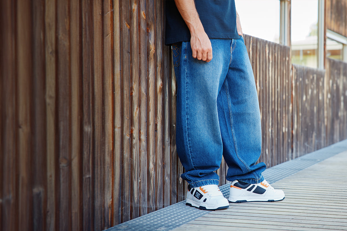 A person wearing loose blue jeans, a dark navy t-shirt, and white sneakers with orange and black accents, standing on a wooden boardwalk next to a wooden fence.