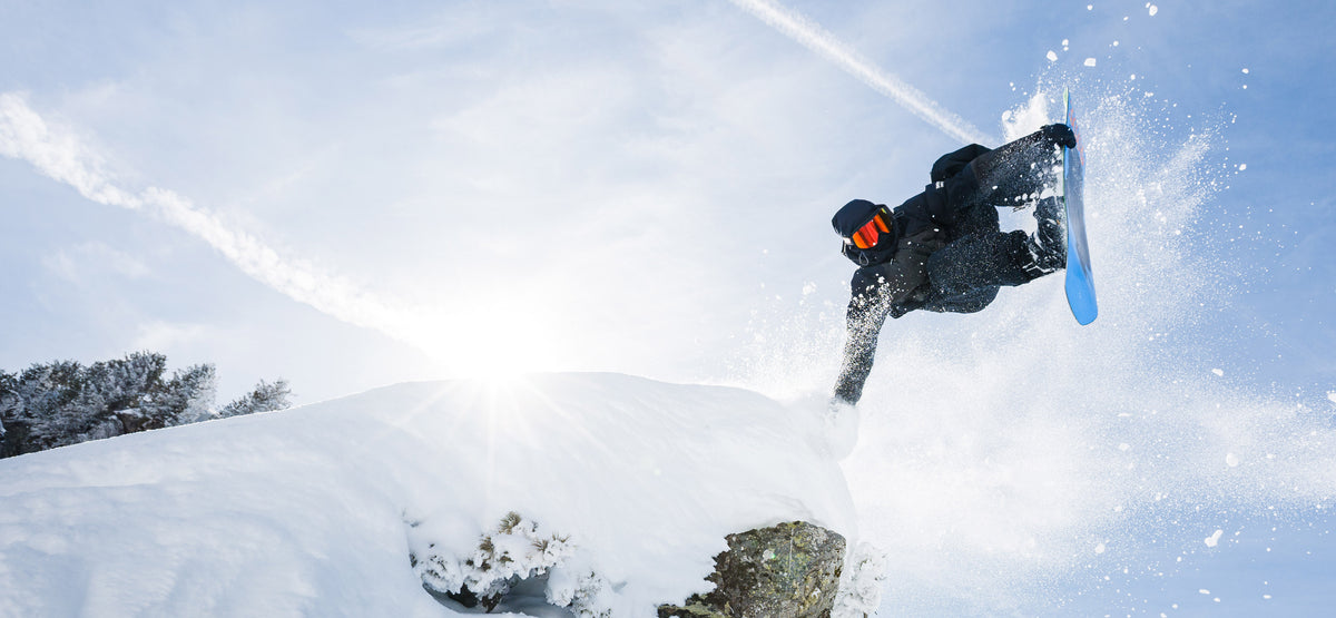 A snowboarder in black gear mid-air performing a jump against a bright snowy sky with snow spraying around them.