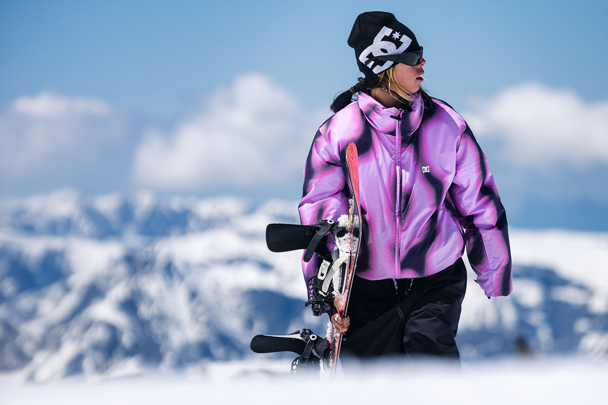 A skier wearing a purple and black patterned ski jacket, black pants, and a black beanie with a white logo walks through the snow carrying ski equipment, with a blurred mountain background under a blue sky.