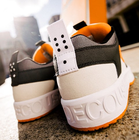 Close-up of the back of two stylish sneakers with orange, white, and gray accents, featuring a white pull tab with black dots and embossed text on the heel, outdoors in a sunny urban setting.