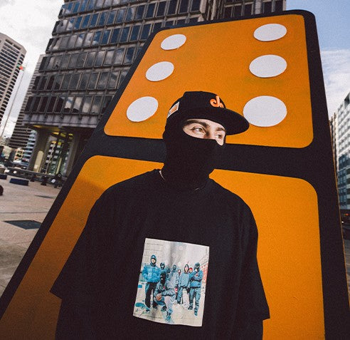 A person in a black mask, cap, and a graphic t-shirt stands in front of a large, upright domino sculpture in an urban setting with modern buildings in the background.