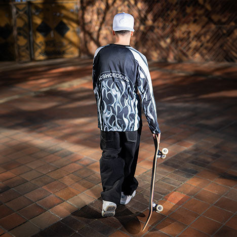 Person in a black shirt with silver chrome details and black pants holding a skateboard on a brick pavement with a textured wall in the background