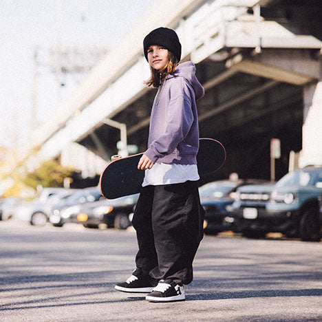 Boy holding a skateboard in an urban setting with cars and a bridge in the background