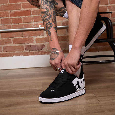 Person tying a black and white sneaker on a wooden floor with a brick wall background