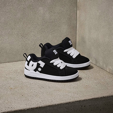 A pair of black and white DC brand sneakers with thick white laces are displayed on a concrete surface against a plain, light-colored background.