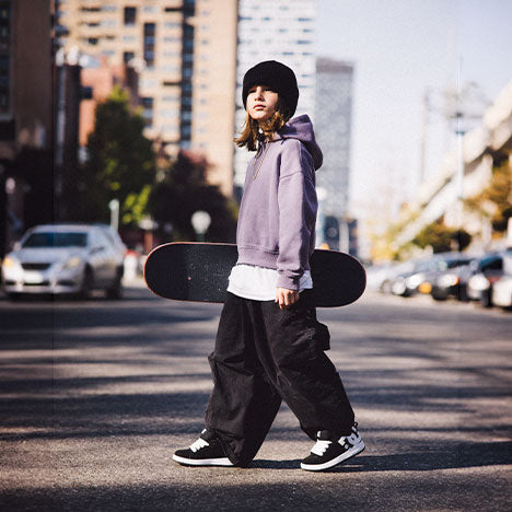 Child in beanie, purple hoodie, black pants, and black and white sneakers holding skateboard while walking outside