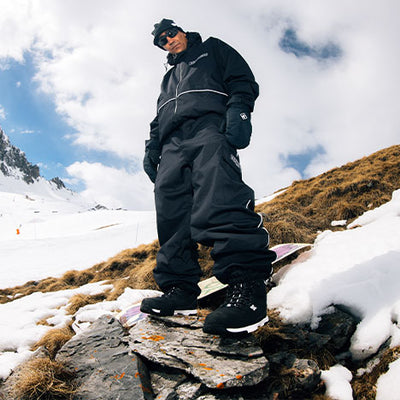 Person in black snowsuit standing on a rocky outcrop with snowy landscape and mountains in the background