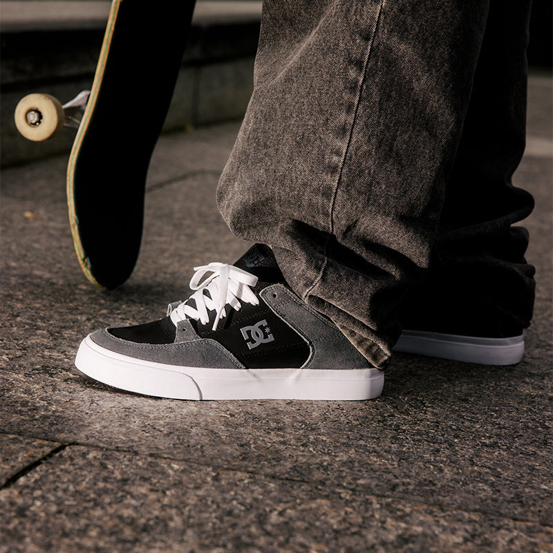 Person wearing black and gray sneakers with a white sole on a concrete surface, with a skateboard in the background.