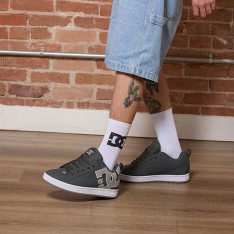 Person wearing gray sneakers with a logo, white socks with a black logo, and denim shorts against a brick wall.