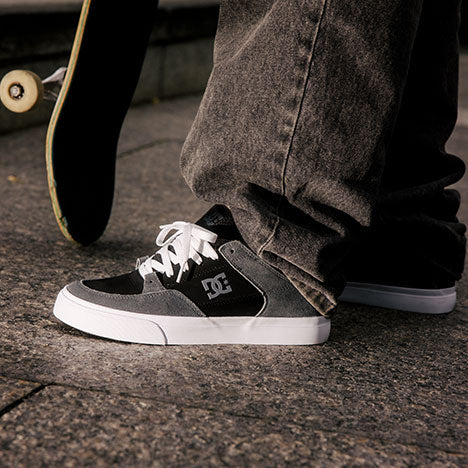 A close-up of a person in gray jeans wearing black and gray DC skate shoes, standing on pavement next to a skateboard.