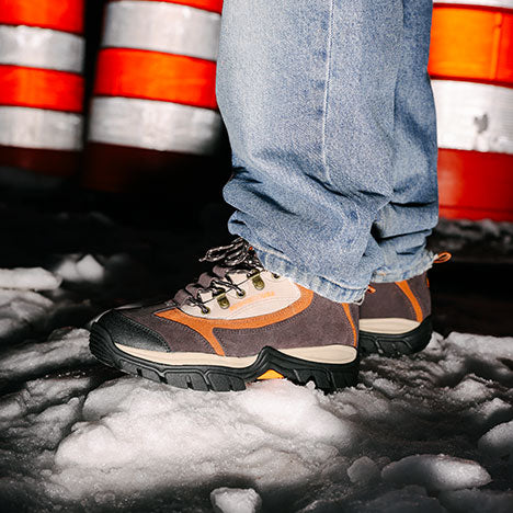 Person wearing black and brown work boots with tan and orange accents and jeans standing on a snowy ground with traffic barrels in the background.
