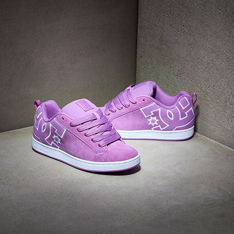 A pair of pink DC brand sneakers with white soles and prominent white DC logos on the sides, displayed against a minimalist grey background.