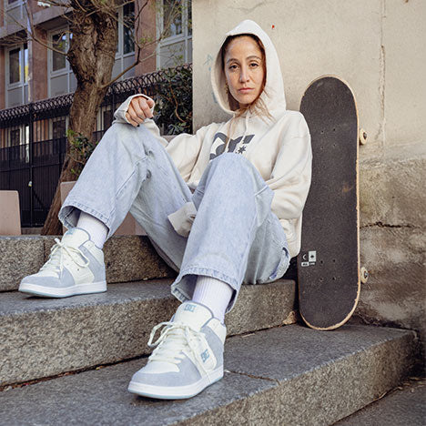 Woman sitting on steps with a skateboard, wearing a hoodie, jeans, and a pair of light blue and white DC sneakers.