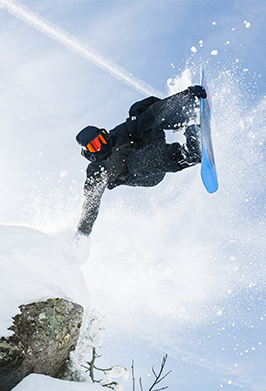 A snowboarder in black gear mid-air performing a jump against a bright snowy sky with snow spraying around them.
