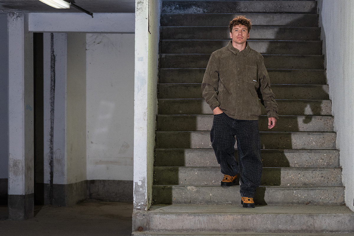 A man wearing a brown corduroy jacket, dark patterned pants, and brown shoes walks down concrete stairs in a dimly lit indoor setting.