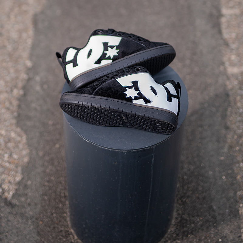A pair of black DC Shoes low-top sneakers with white DC logos and star details, displayed on a dark cylindrical pedestal against an asphalt background.