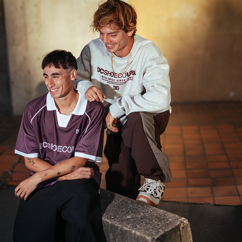 Two young men posing in streetwear. The seated man wears a maroon and white DOSHECOURS jersey, while the standing man wears a light gray sweatshirt with a dark graphic and dark brown pants.