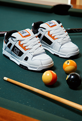 White, black, and orange DC sneakers displayed on a green pool table beside billiard balls and a cue.