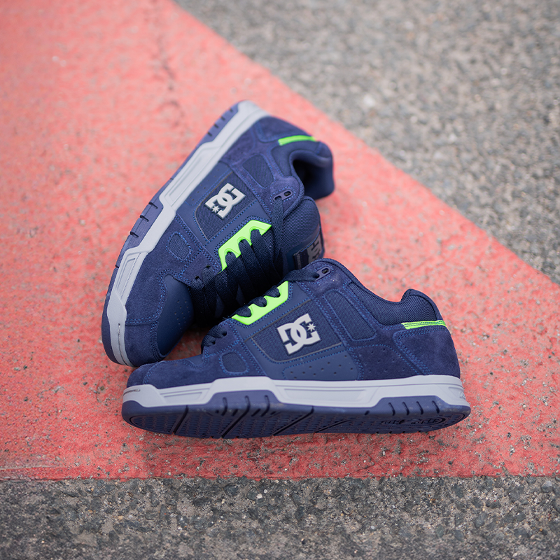 A pair of DC skate shoes in navy blue suede with bright neon green accents and white soles, resting on a red painted line of asphalt.