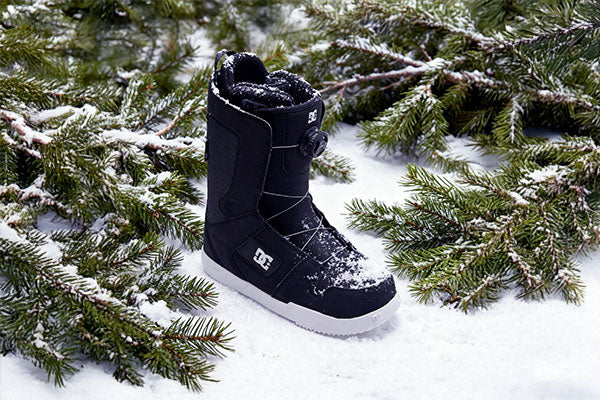 Black snow boot with white sole on a snowy ground with pine branches.
