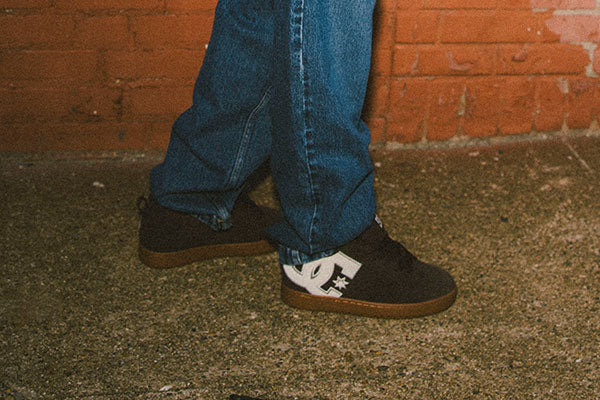 Person wearing black and white DC Shoes sneakers and blue jeans against a brick wall.