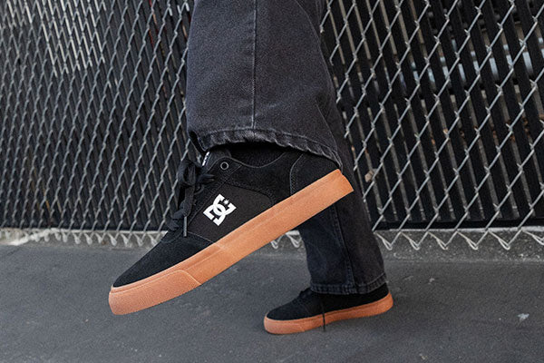 Black DC shoes with gum soles worn by a person against a chain-link fence background