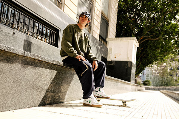 Person sitting on a bench with a skateboard, wearing a cap and casual clothing, in an urban setting.