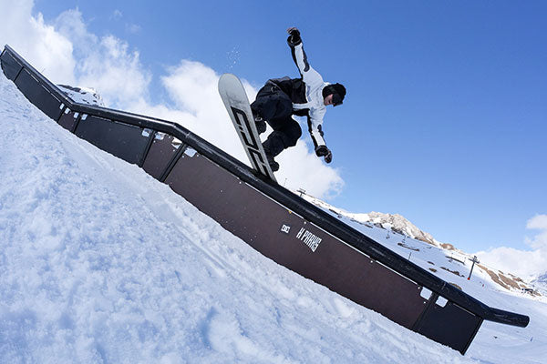 Person snowboarding off a rail with a snowy landscape and blue sky in the background