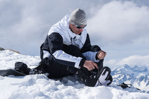 Person with beanie and sunglasses sitting on a snowy mountain adjusting their snowboard boot.