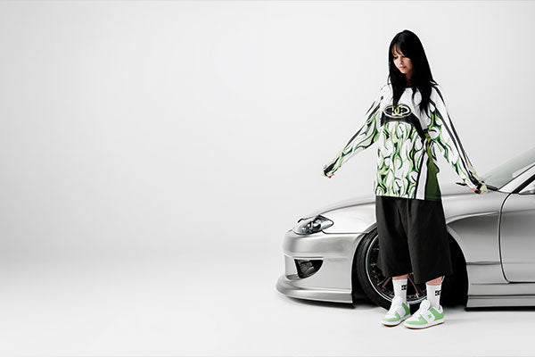 Person wearing a green and white patterned outfit standing next to a silver car on a white background