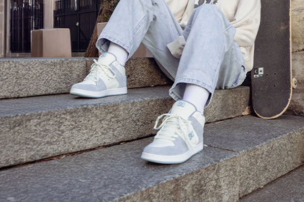 Woman sitting on steps wearing light blue and white Manteca sneakers with a skateboard next to her.