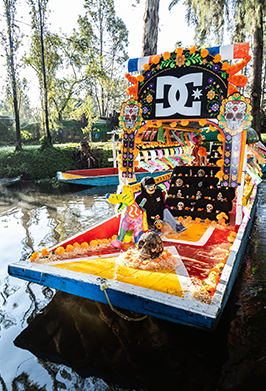 A colorful, decorated DC-branded boat floats on a canal during what appears to be a festive outdoor event. The boat features a large DC logo on the back, bright orange, yellow, red, and blue painted sections, floral decorations, and a person standing near the center.