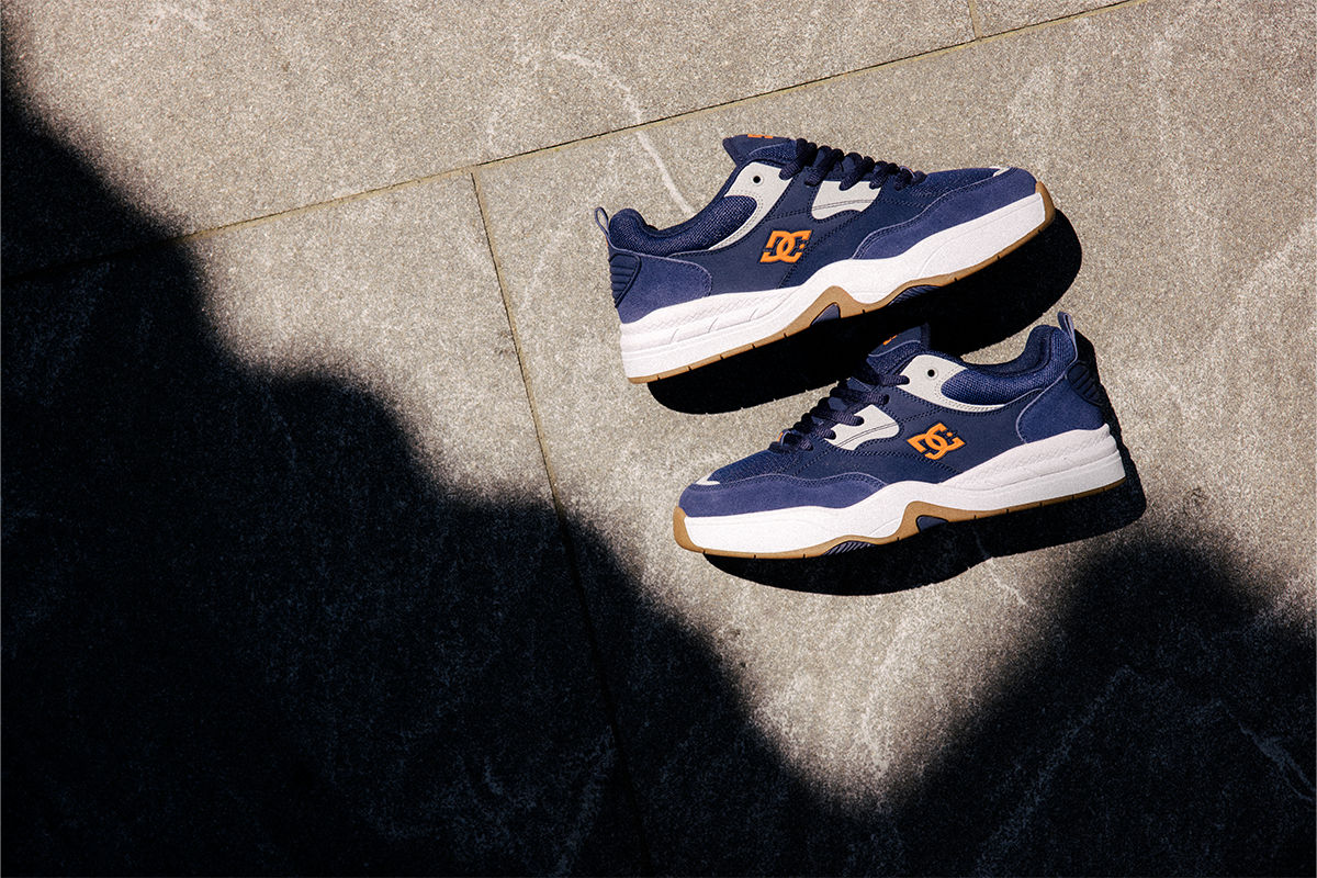 A pair of DC sneakers in navy blue, white, and gum brown, displayed on a gray stone tile floor under direct sunlight with a prominent shadow.