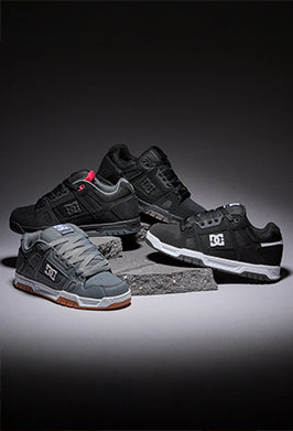 Four DC sneakers arranged on stacked stone blocks against a dark studio background. The set includes black and dark gray models with white soles, plus one gray model with a brown gum sole.