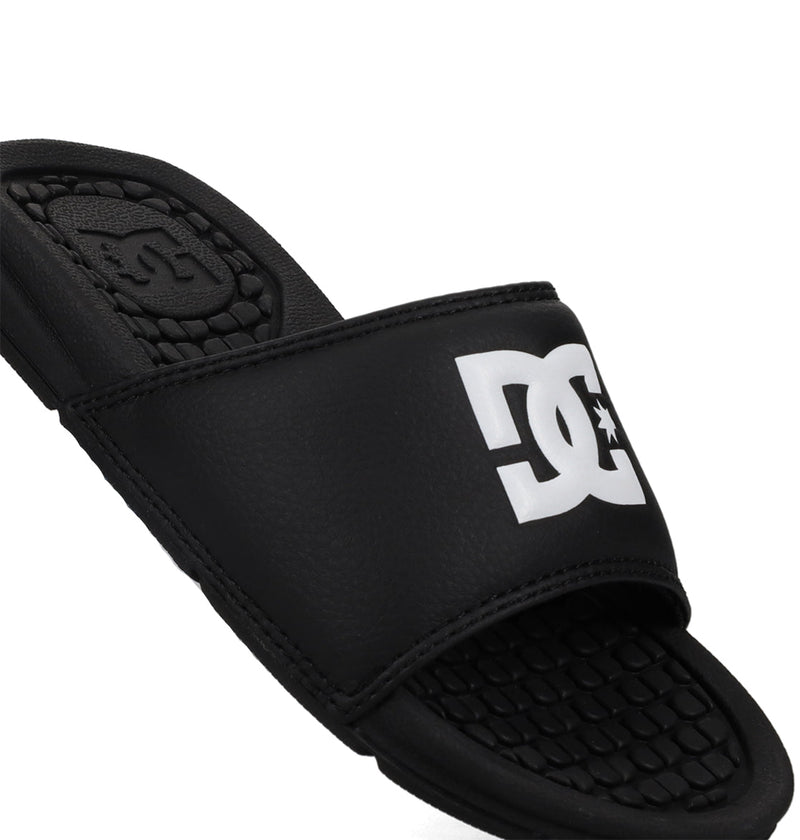 A close-up of the Kids' Bolsa Slides by DC Shoes shows a black sandal with a white DC logo on the strap and a textured footbed for comfort.