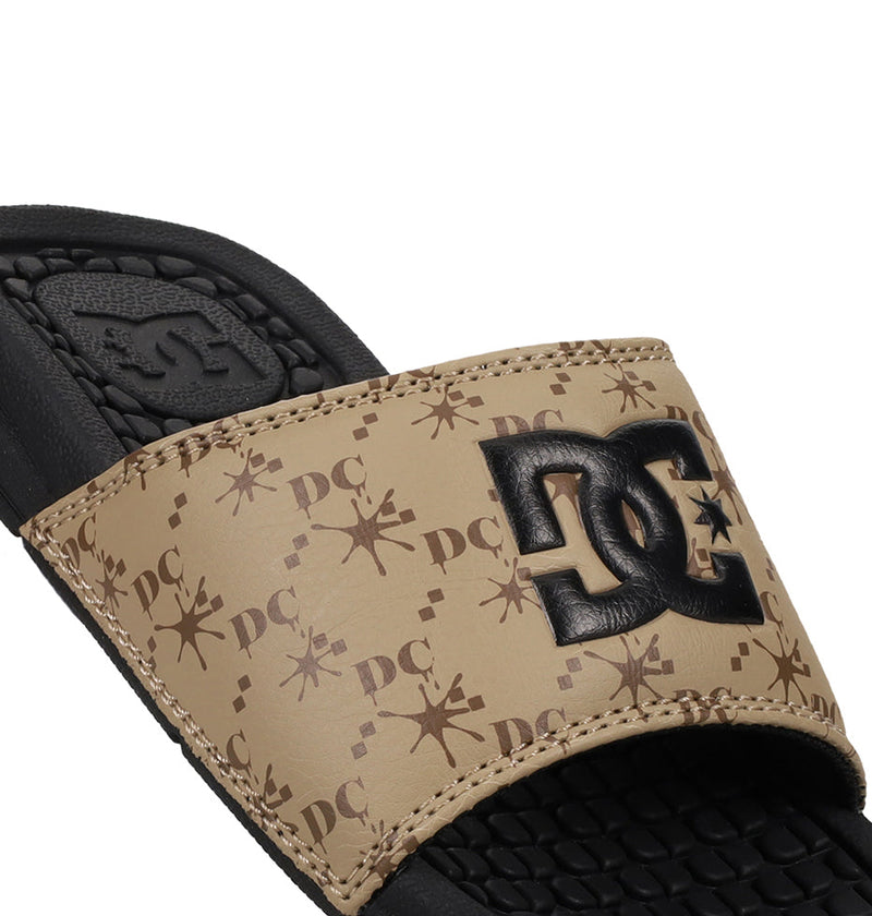 A close-up of the DC Shoes Kids' Bolsa Slides shows a tan strap with repeating DC and star patterns and a bold black DC logo. The textured black footbed adds comfort, making them perfect as kids' slide sandals.