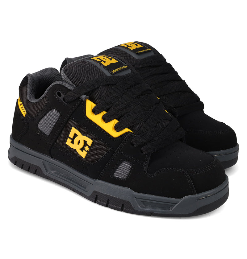 A pair of DC Shoes Men's Stag skate shoes in black with yellow accents, logo, thick black laces, grey soles, padded tongue, and durable cupsole—ideal for skate culture fans—displayed side by side on a white background.