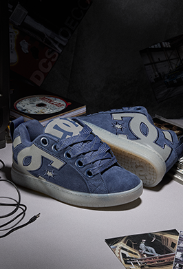 A pair of DC Comics low-top sneakers in blue suede and canvas, featuring a large white DC logo on the side and a translucent white sole, displayed on a tabletop with a dark background.