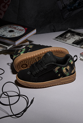 A pair of DC Shoes low-top sneakers with black uppers, green camouflage side panels, and tan gum soles, displayed on a tabletop with books and cables in the background.