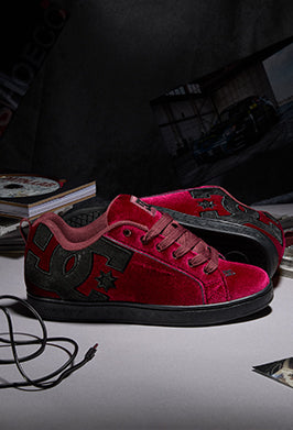 Red and black DC sneakers with a velvet texture, displayed on a dark surface with a studio background.