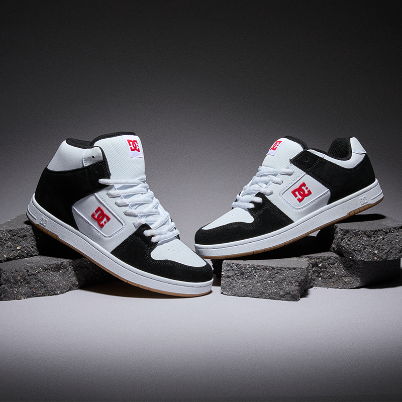A pair of DC brand mid-top sneakers in black, white, and red, styled on dark gray textured blocks against a dark gray studio background.