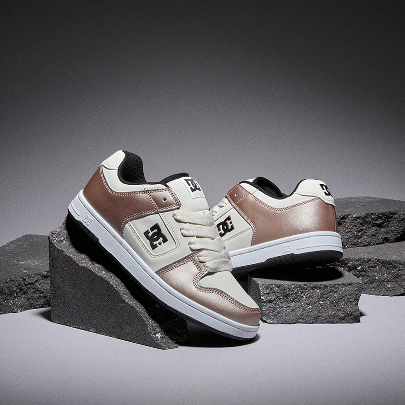 A pair of DC skate shoes in metallic rose gold and off-white, styled on dark gray concrete blocks against a dark gray studio background.